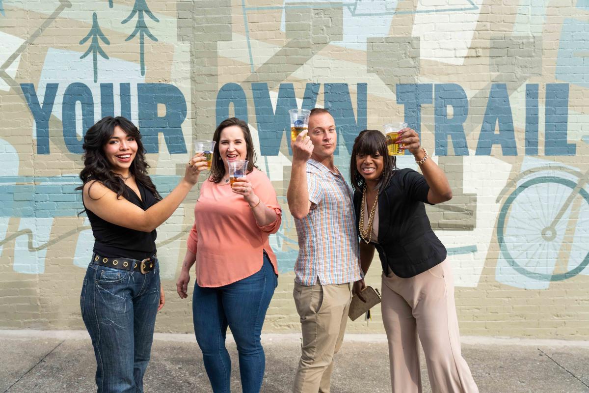 Four people are standing in front of a mural holding their hub cups in The Hub in Downtown Elizabethtown