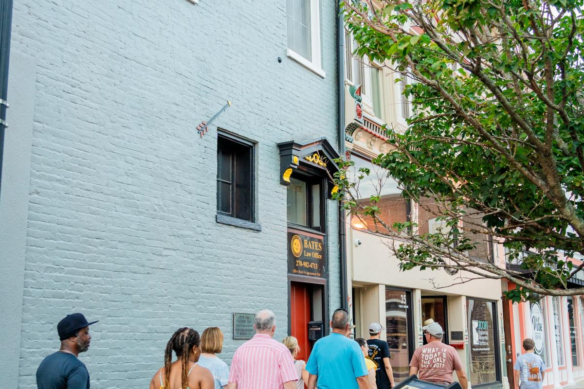 A group of people walking along a sidewalk in a charming downtown area, passing by historic buildings with a tree and informational plaque in the foreground