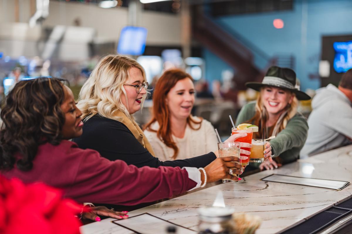 Four friends laughing and clinking their drinks together at Social on Main, enjoying a casual and cheerful moment at the bar.