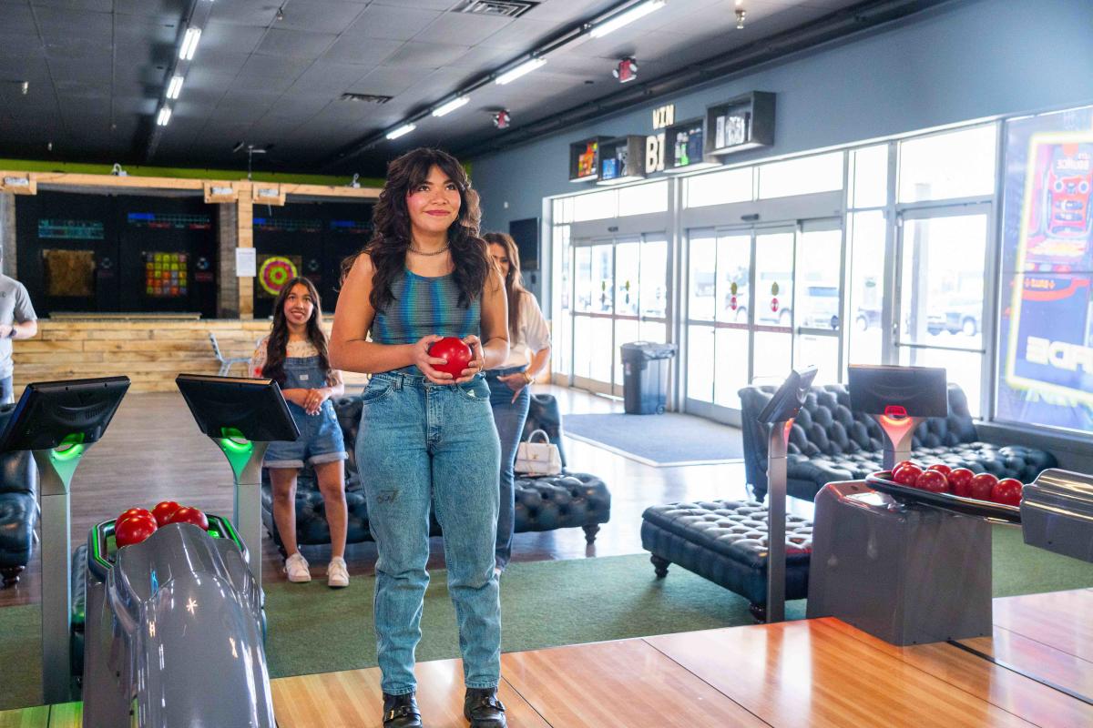 A young woman holds a bowling ball, smiling as she stands on a bowling lane.