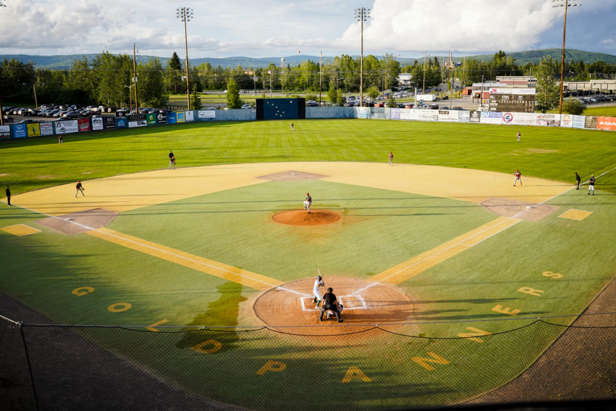 Wide view of the Midnight Sun Baseball Game, with players positioned across the field under bright evening sunlight and mountains in the background.