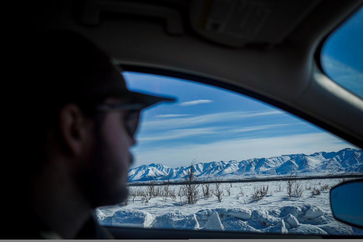 A person driving a car through a snowy landscape with clear skies and mountains in the distance. The focus is on the view outside the window, highlighting the scenic winter drive.