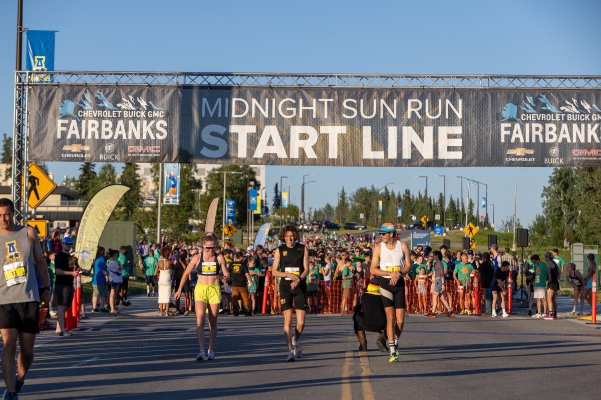 Runners and spectators gather beneath the Midnight Sun Run start line banner in Fairbanks, Alaska, during a lively summer race under bright evening daylight.