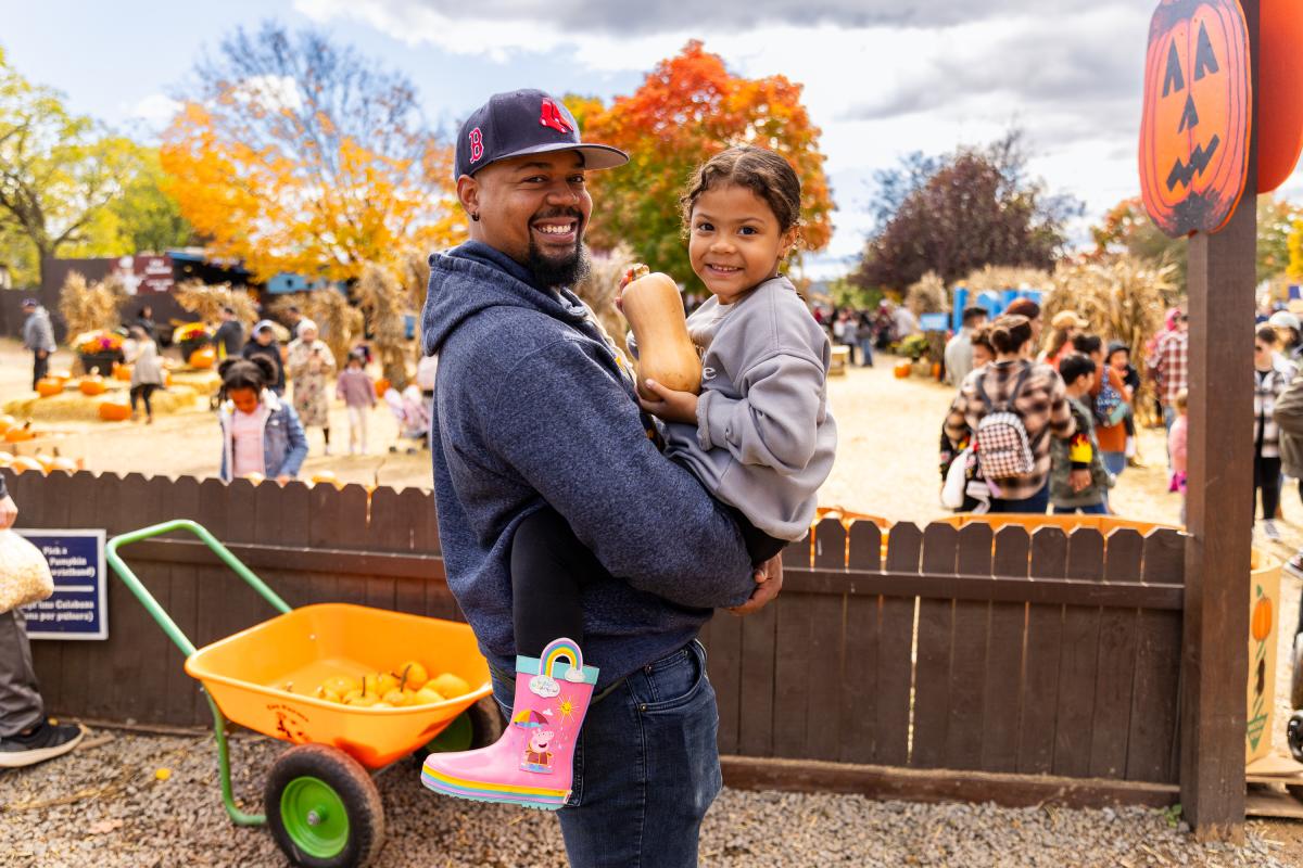 A man holds a smiling girl in a pumpkin patch, surrounded by autumn foliage and families enjoying the festive atmosphere. A wheelbarrow filled with pumpkins is nearby.