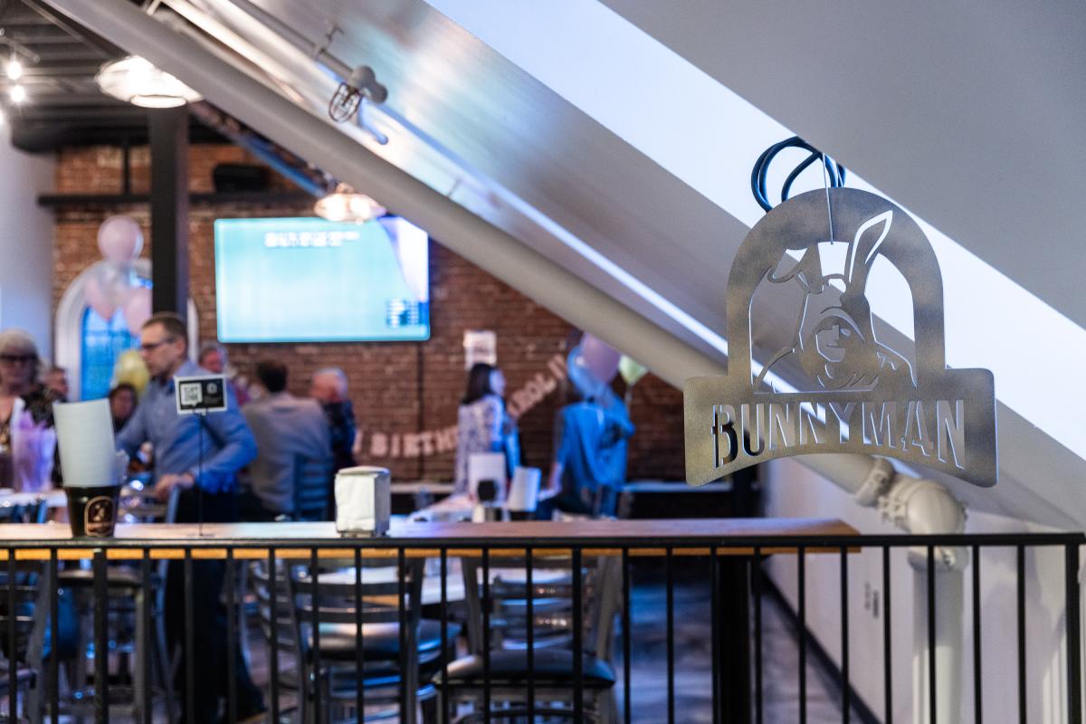 Metal sign reading "BUNNY MAN" hangs above a bustling bar area with patrons socializing, exposed brick walls, and modern decor.