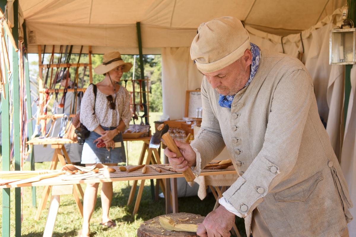 A craftsman in period attire carves wood with a hammer, while a woman observes nearby in a tented market setting. Tools and crafts are displayed.