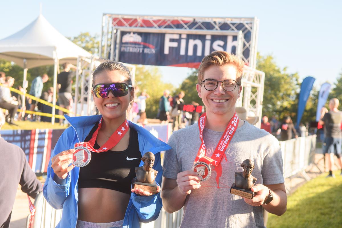 Two smiling participants hold trophies and medals at a finish line, with a "Finish" banner in the background and spectators nearby