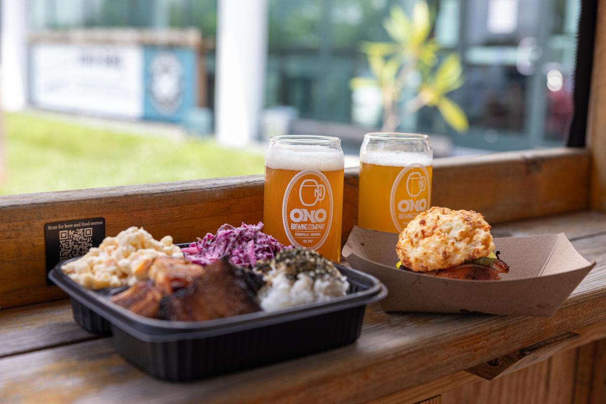 A wooden table displays a takeout box with barbecue, sides, and two glasses of beer, set against a bright outdoor backdrop.