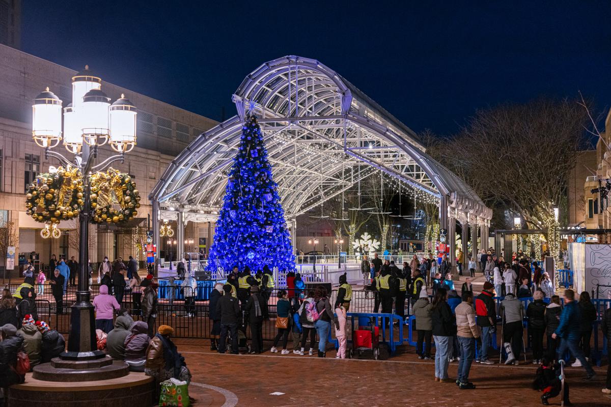 A festive outdoor plaza at night, featuring a large blue Christmas tree under a glass canopy, surrounded by crowds and holiday decorations.