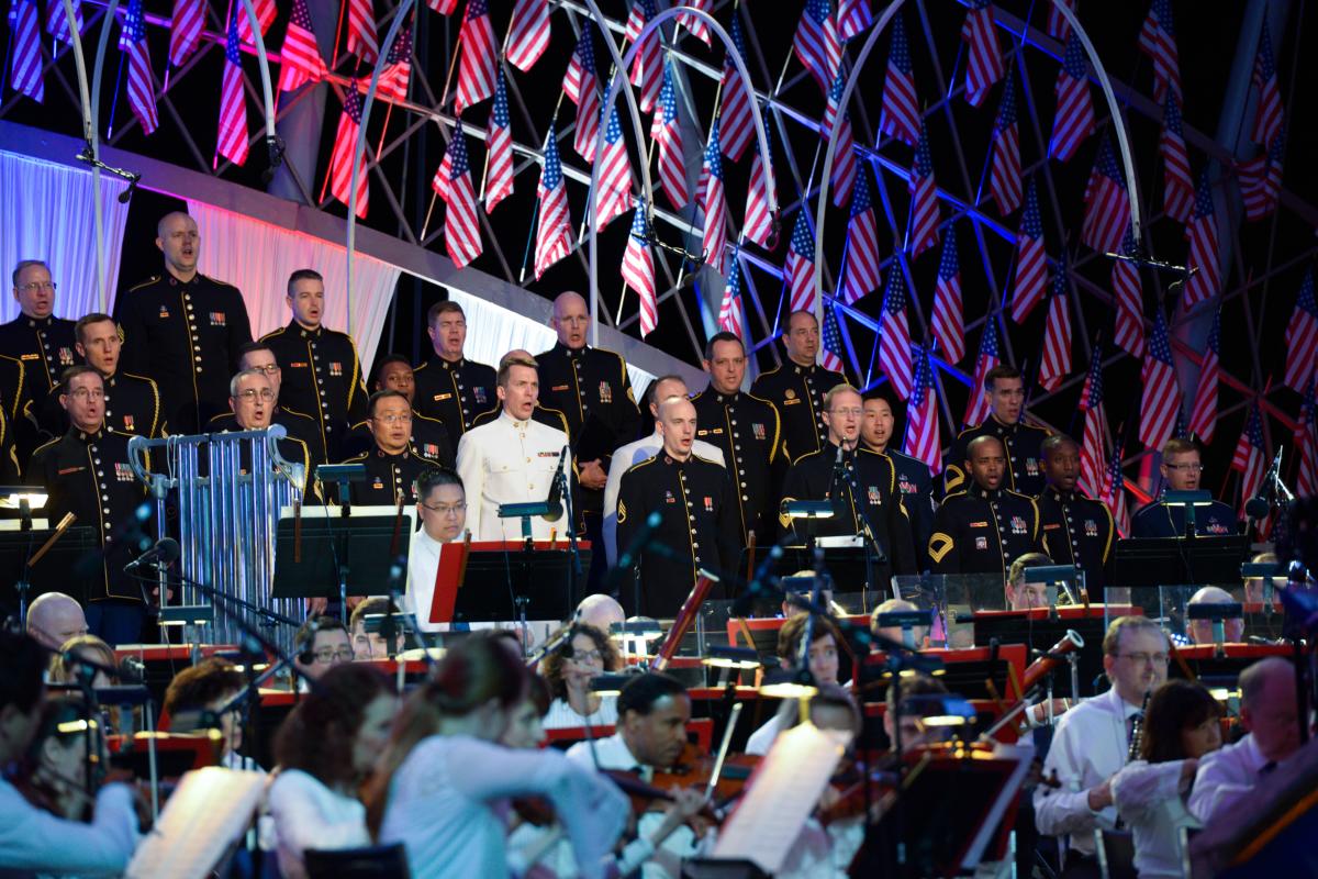 National Memorial Day Concert of choir singing patriotic tunes flanked by american flags and a symphony orchestra