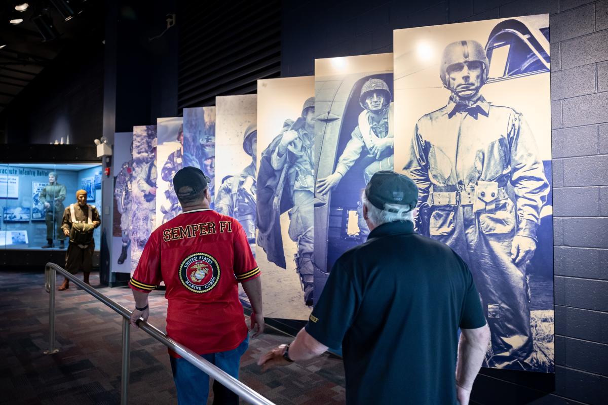 Two visitors walk through a military history exhibit lined with large blue-toned panels of paratroopers and nearby display cases at Fayetteville's ASOM.