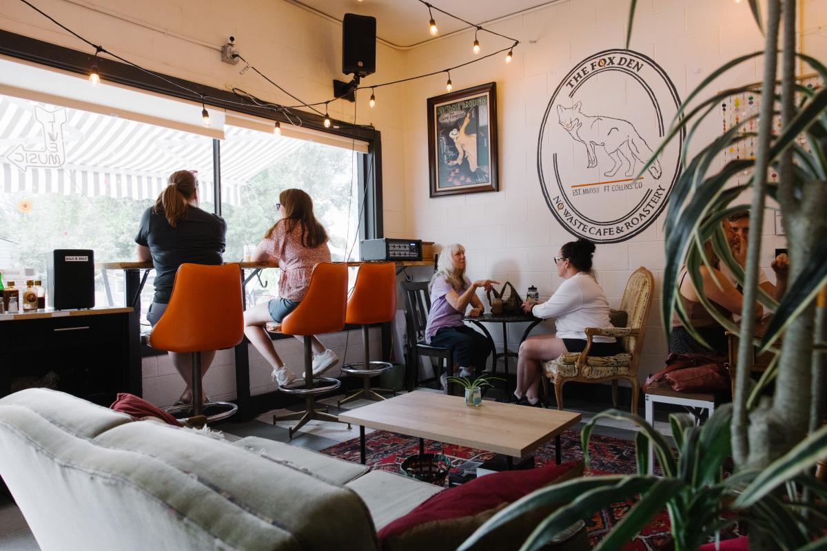 Four people sitting in a coffee shop with a mural of a fox on the wall that says The Fox Den, No Waste Cafe and Roastery