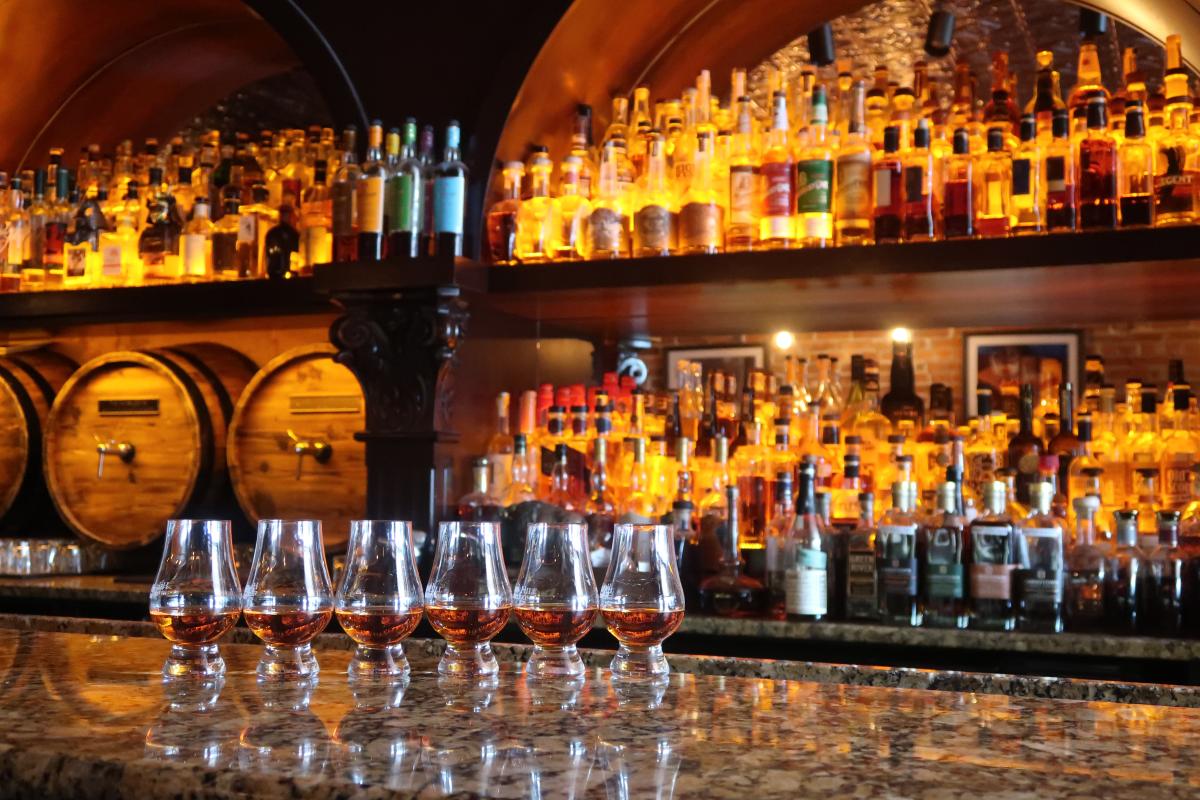 Six tasters of whiskey in front of a wall filled with whiskey barrels and bottles
