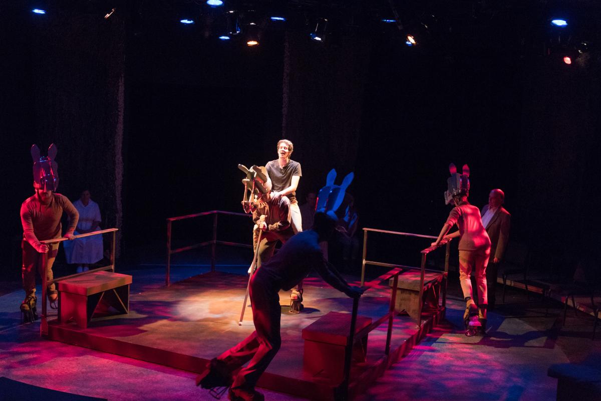 People in horse costumes on the sides of the stage with a person riding a wooden horse in the middle of the stage