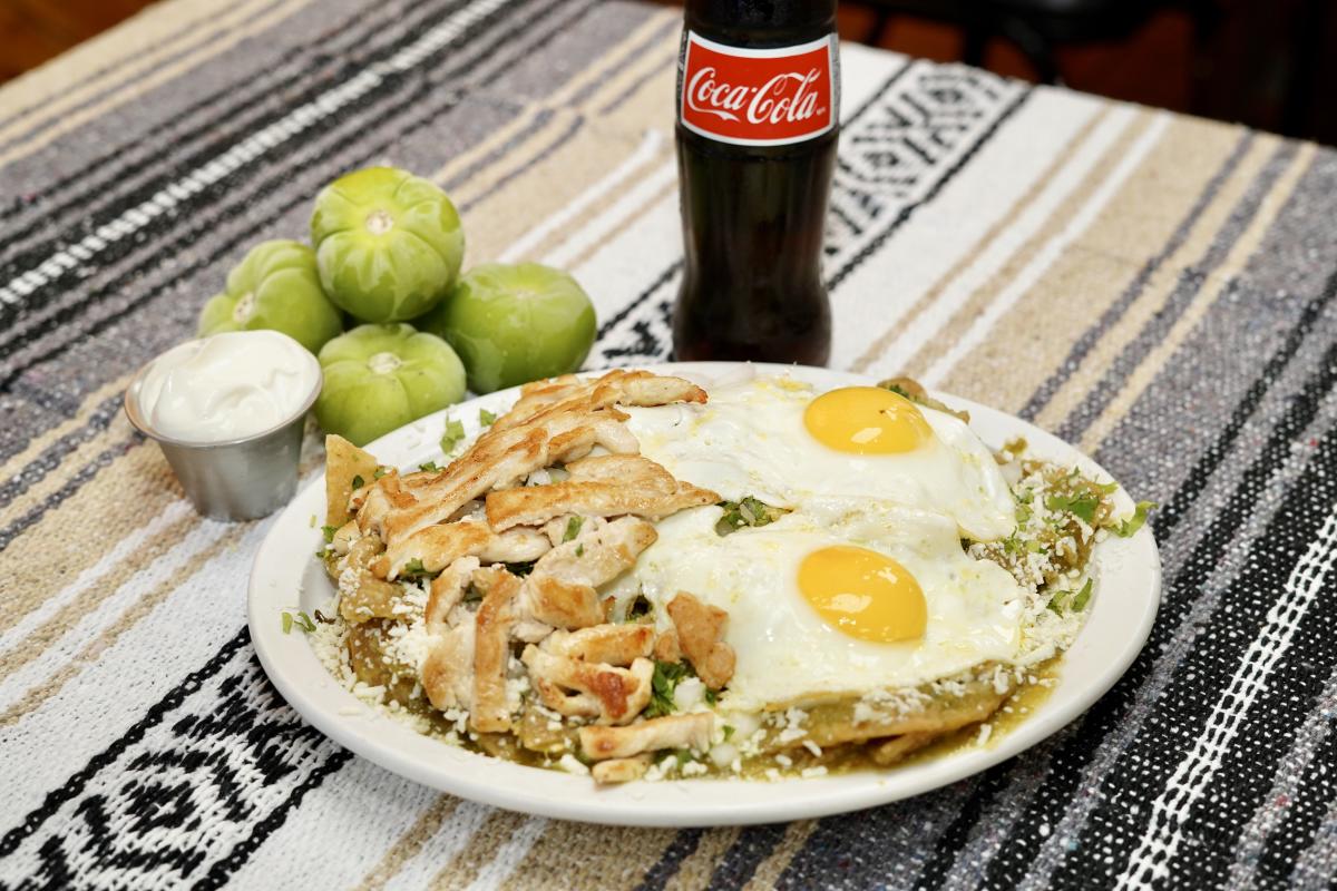 A breakfast dish with chicken, eggs, a green sauce, cheese and tortillas is on a plate in the center of the picture. There is a Mexican Coke, a white sauce, and various vegetables on the side of the plate.