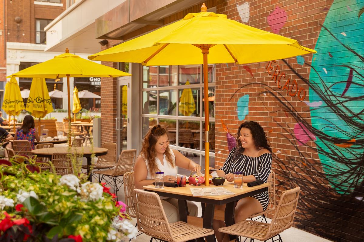 two women eating at an outdoor table at a restaurant sitting under a yellow umbrella with a mural on the wall behind them