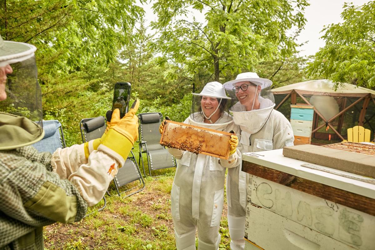A person taking a picture of two people holding a bee hive, they're all wearing beekeeping suits.