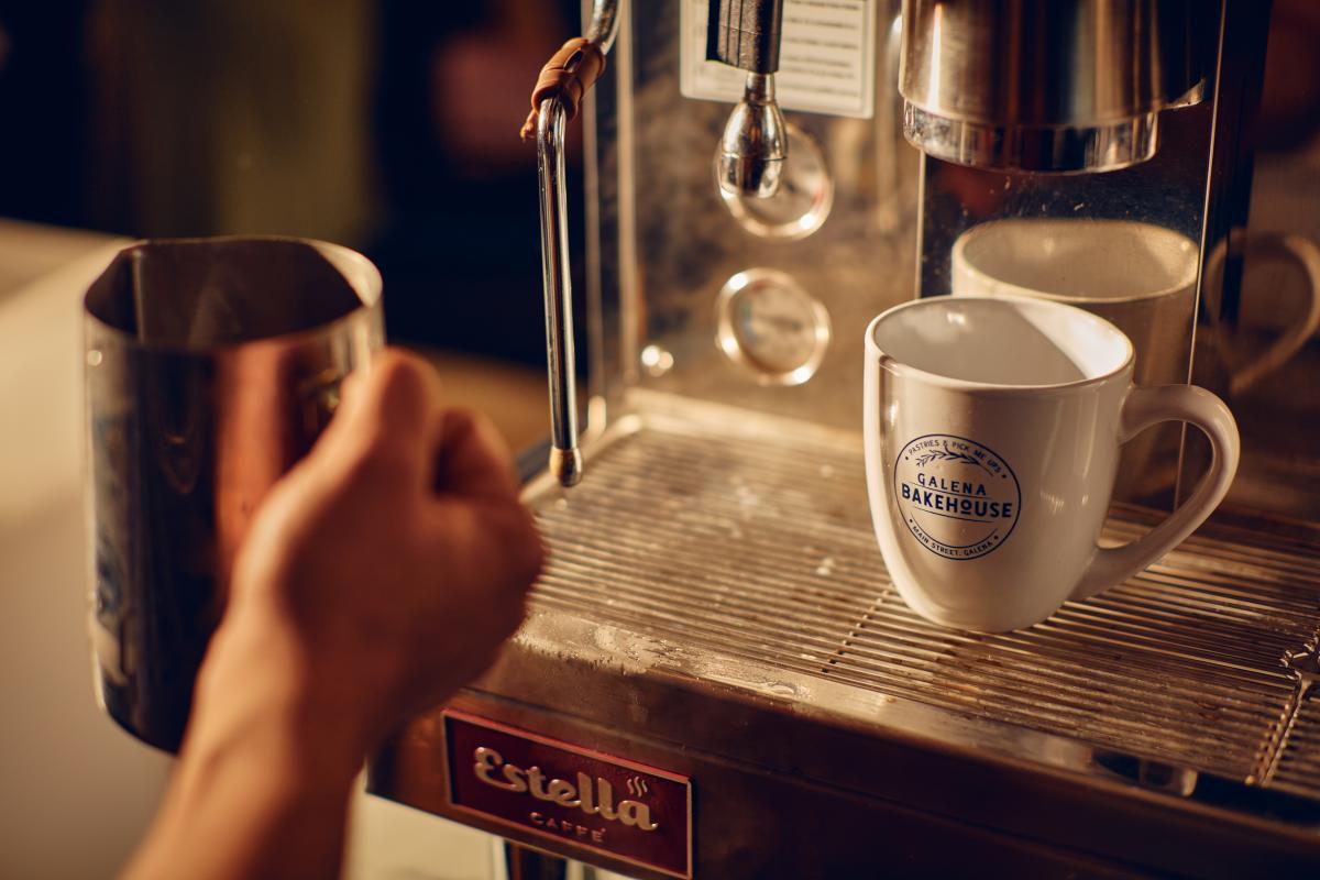 A hand holding a steamer cup in front of an espresso machine, with a coffee cup that says, Galena Bakehouse.