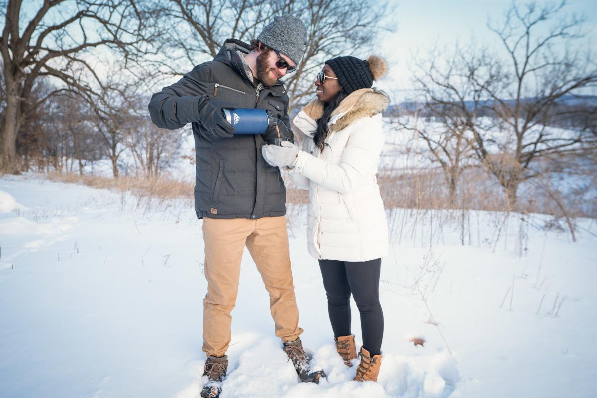 A couple standing in the snow, sharing a hot beverage. They are wearing winter clothes, like hats, boots, and coats.