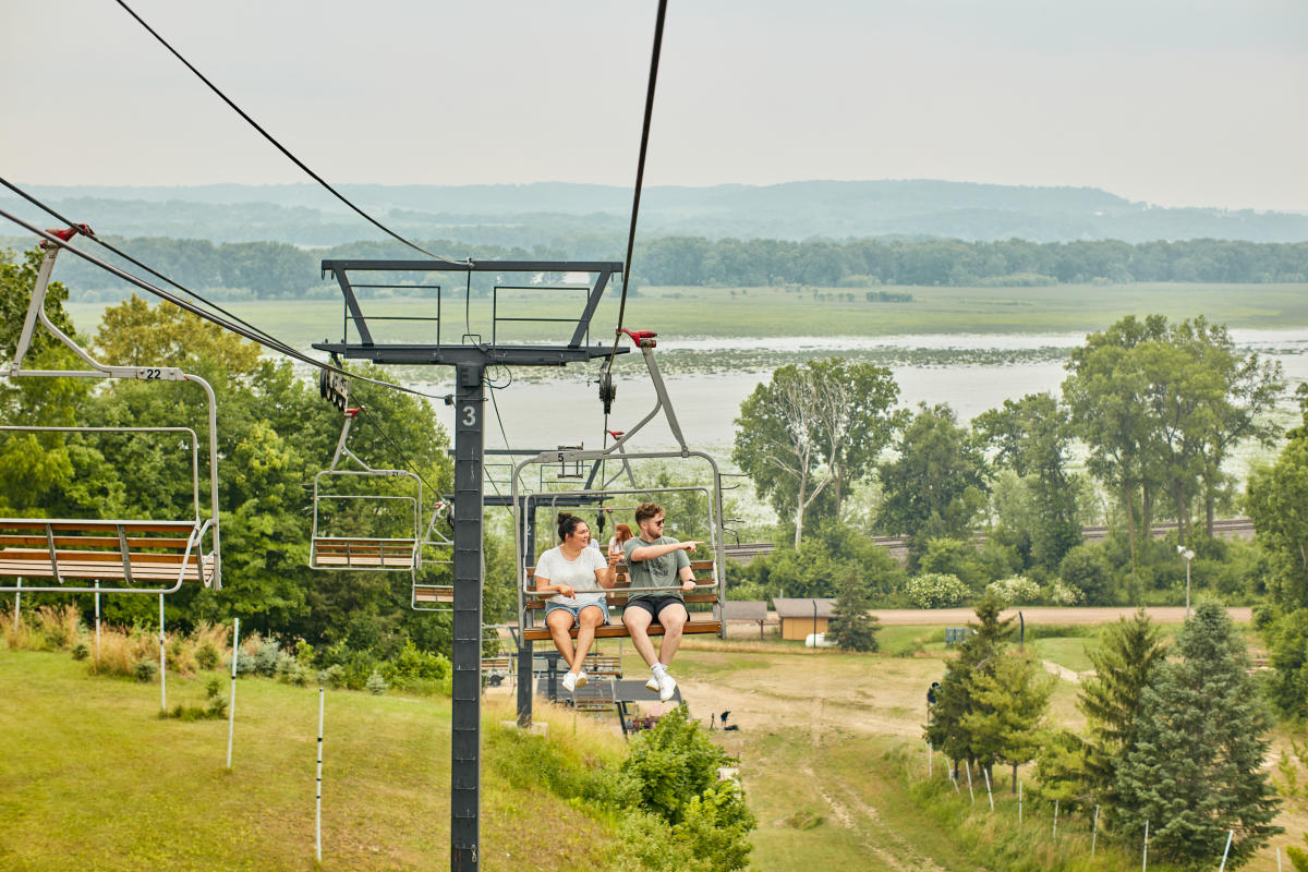 Two people riding a chairlift in the summer, with the Mississippi River behind them.