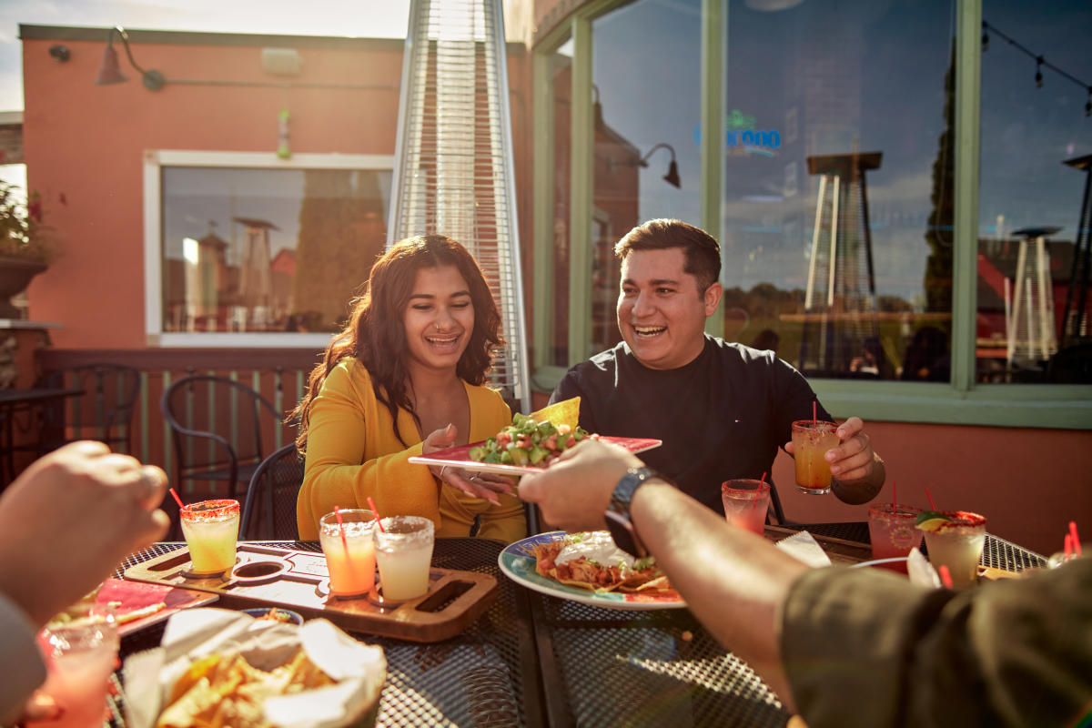Four people sitting at an outdoor patio table enjoying Mexican food. One person is handing another a plate of food.