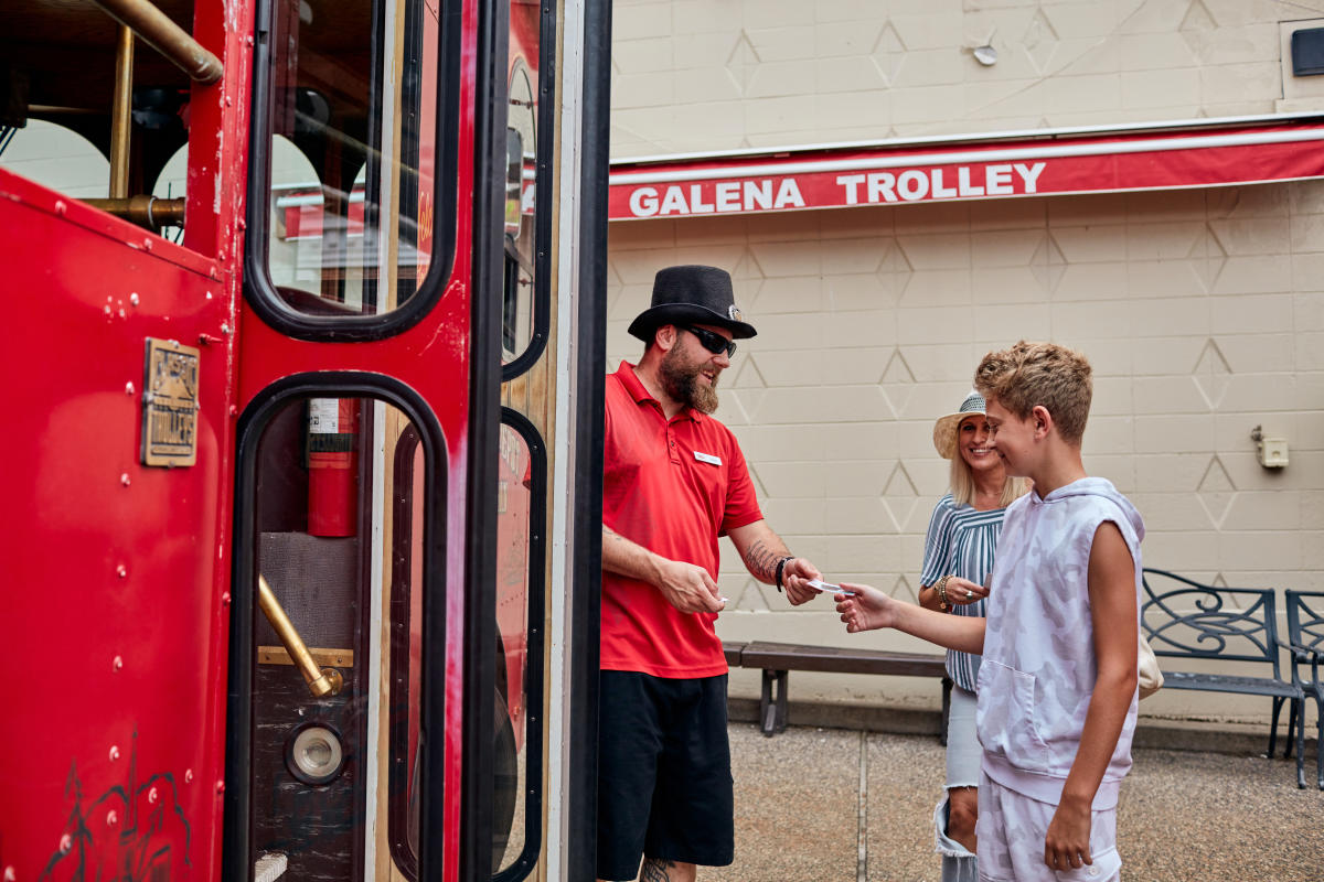 Three people standing next to a red trolley. A child is handing a ticket to an employee while their mother is waiting to board the trolley.