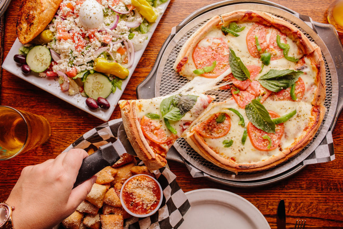 Pizza, a salad, and bread bites on a table. A person serving a slice of pizza, picking it up with a pizza server.