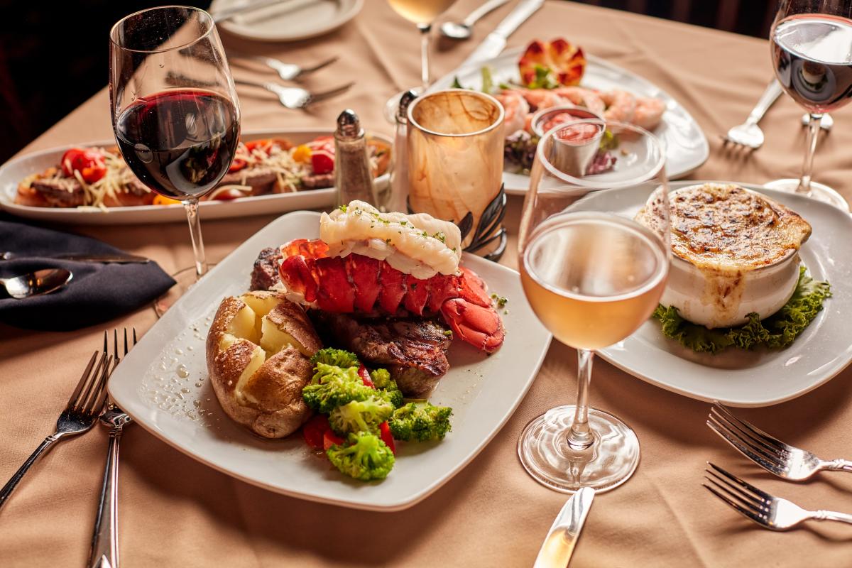 Table of food including lobster tail and baked potato, glasses of wine, French onion soup.