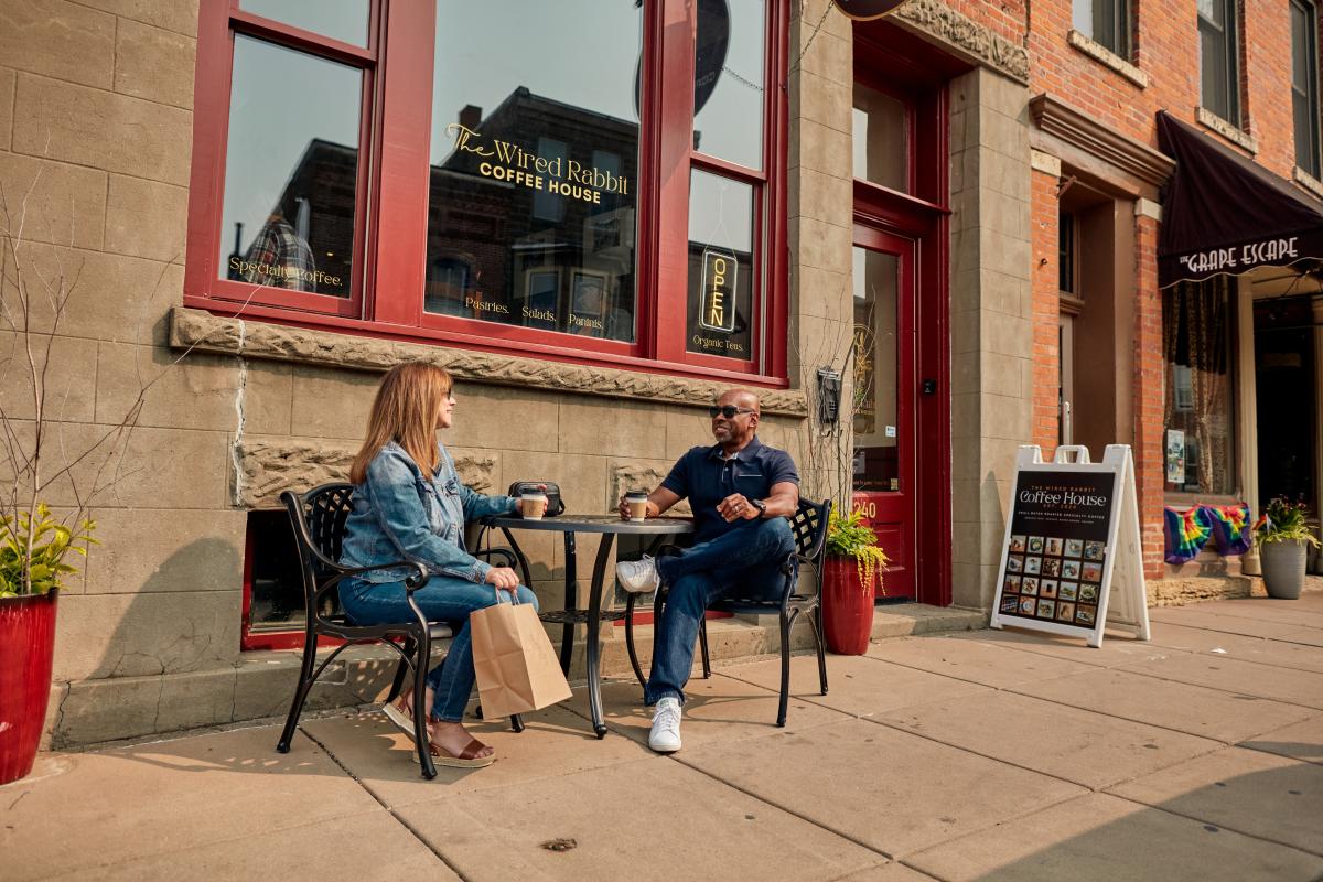 Two people sitting in front of a coffee shop, drinking coffee and smiling.