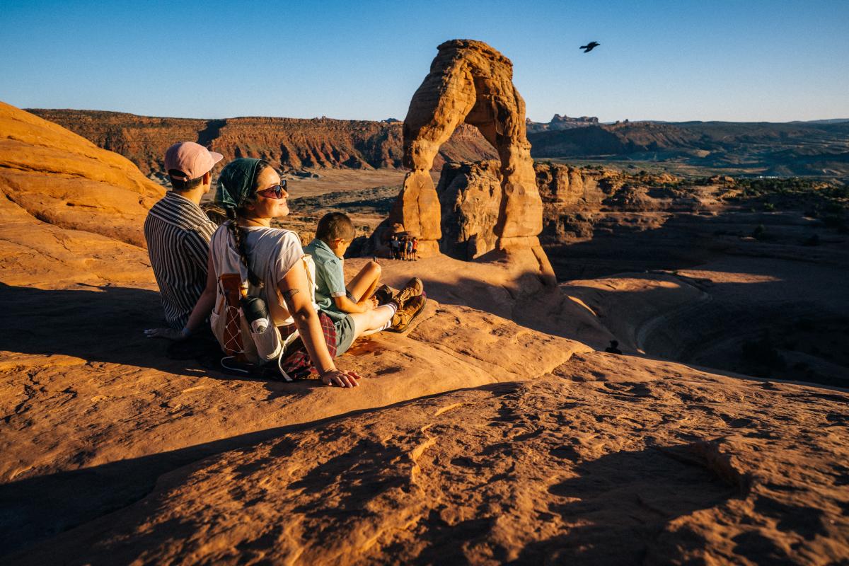 Three people sitting on the red sandstone rocks at sunrise next to the iconic Delicate Arch in Arches National Park, near Moab, Utah