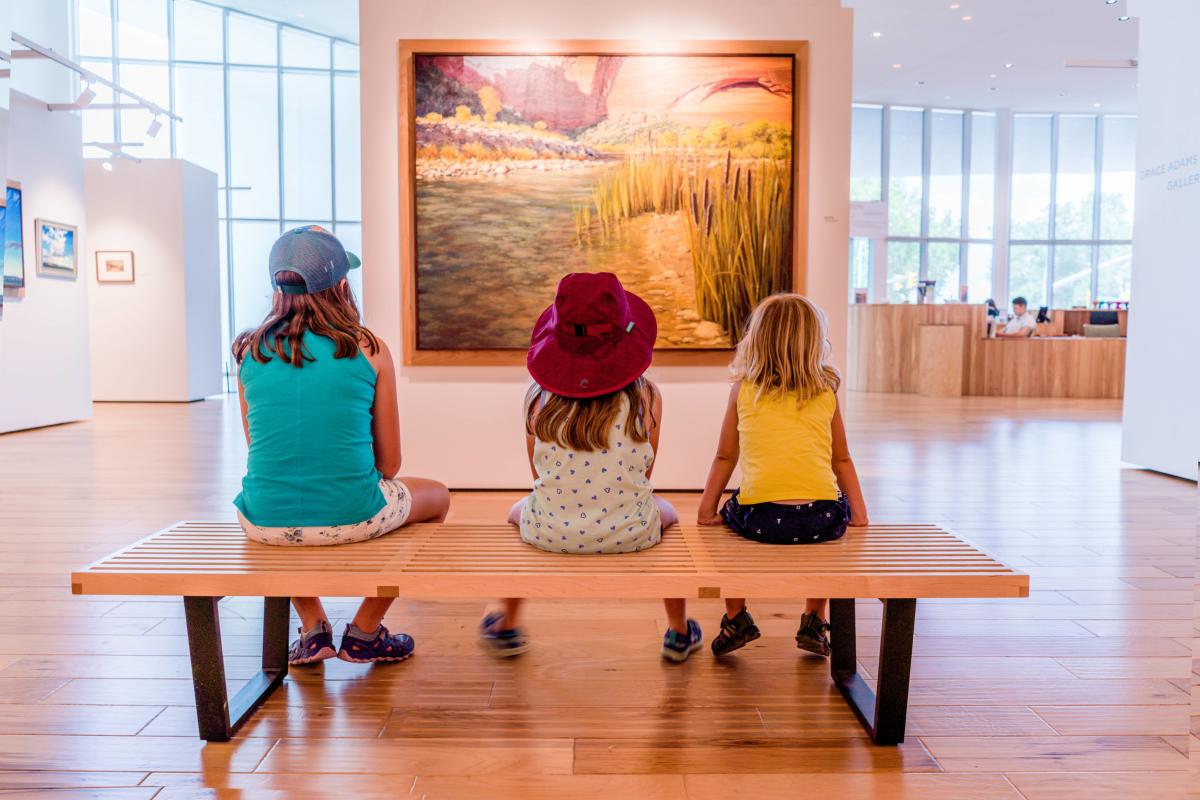 Three kids sitting on a wood bench in front of a landscape painting at the Southern Utah Museum of Art.