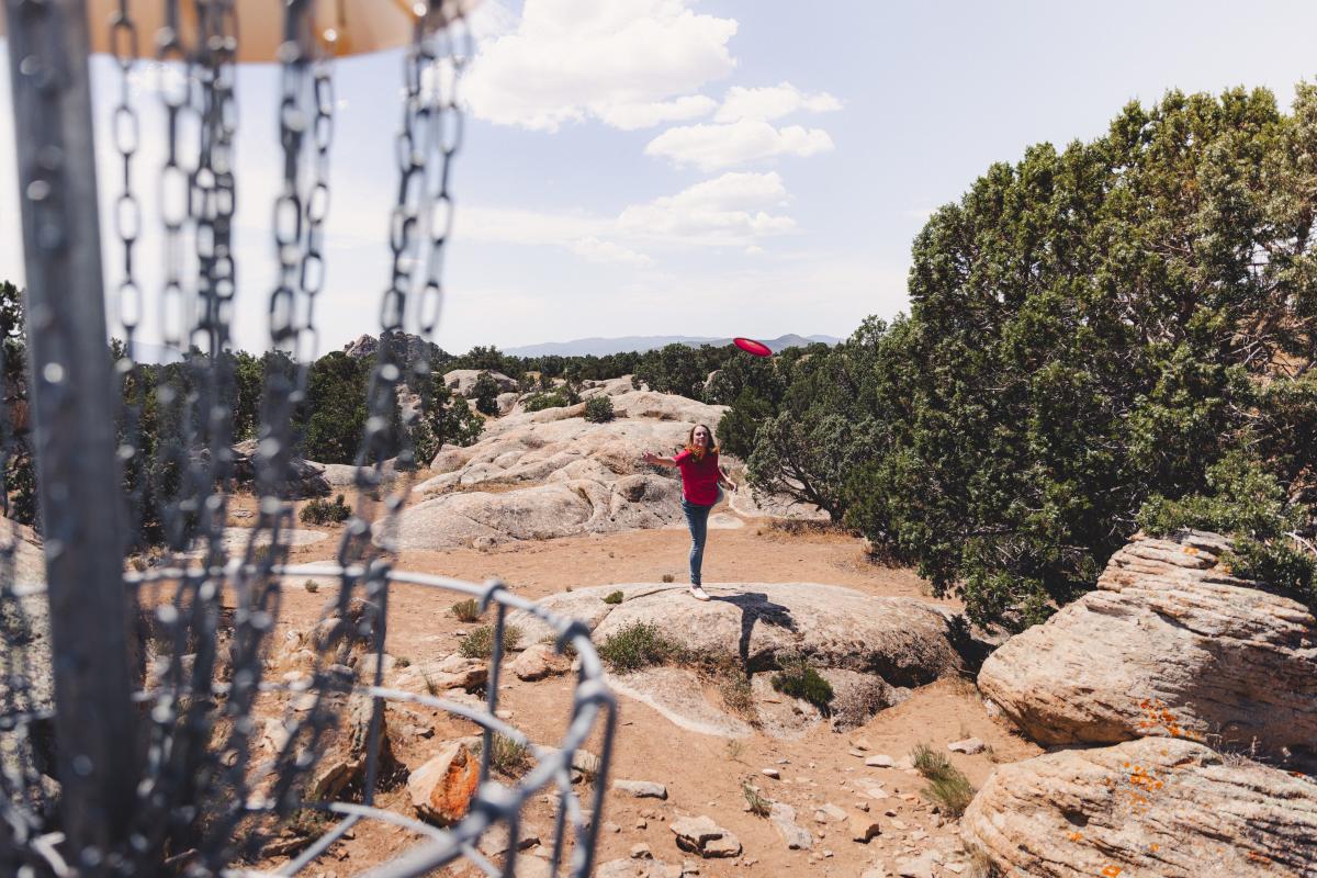 A person dressed in red throwing a disc into a disc golf basket in Three Peaks Recreation Area