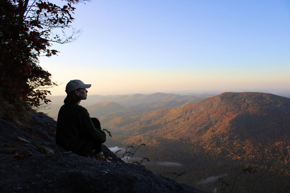 A stunning sunset view from the summit of Whiteside Mountain, North Carolina, featuring a hiker sitting on a rocky edge and gazing out. The extensive range of Blue Ridge Mountains below is illuminated by golden light, showcasing brilliant fall colors (orange, red, gold), with distant hazy peaks and low fog under a gradient sky.
