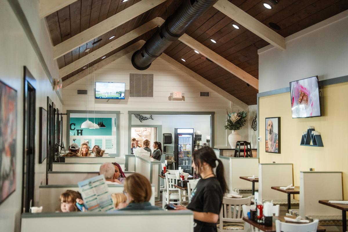 The interior of a bright, modern diner with a high vaulted wood-paneled ceiling and an exposed black air duct. Patrons are seated in white booths throughout the room, and a kitchen service area is visible in the background.