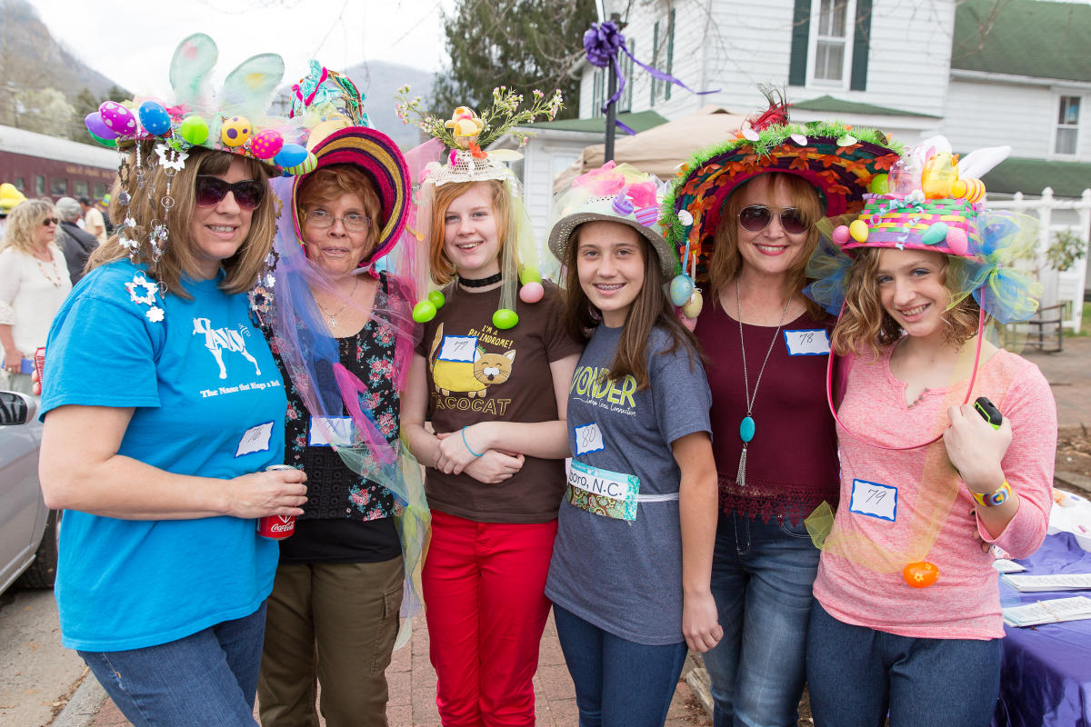 ix women and girls pose together wearing elaborate, whimsical, and colorful handmade Easter hats decorated with bunny ears, plastic eggs, and ribbons. They are wearing contestant numbers for a parade or competition.