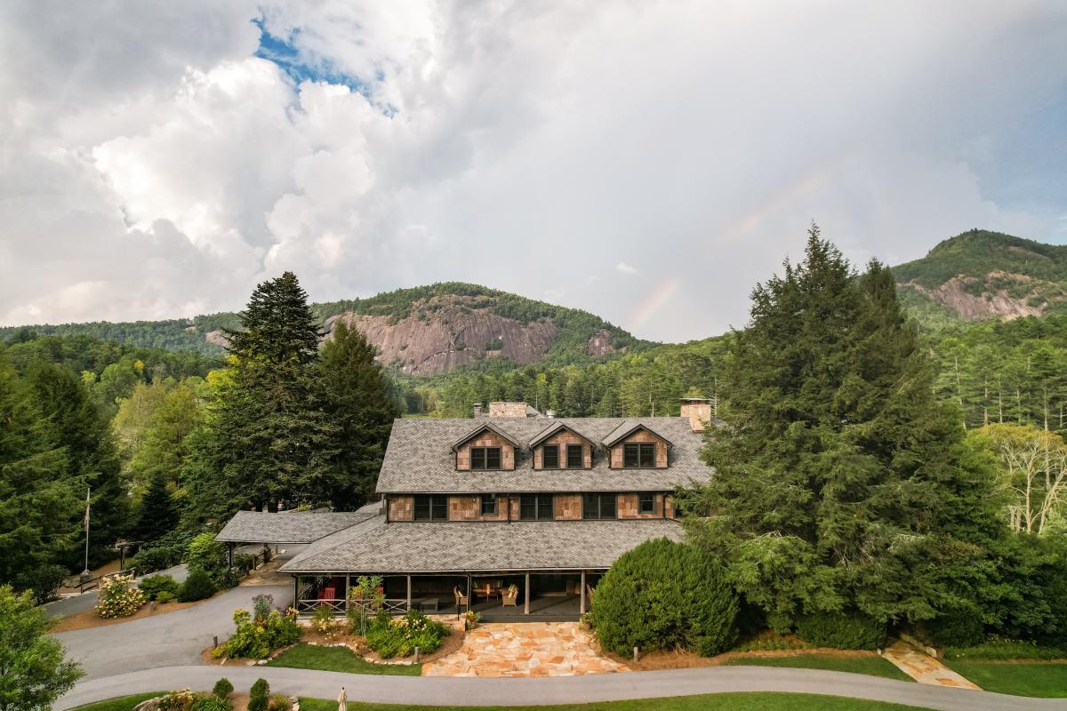 An aerial view of a large, historic mountain inn featuring brown cedar-shake siding and a gray shingled roof. The building is surrounded by lush green forests and overlooks a mountain peak, with a faint rainbow stretching across a dramatic, cloudy sky.