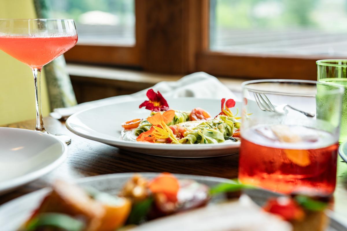 A close-up, shallow-depth-of-field shot of a summer meal, featuring a plate of pasta garnished with bright orange edible flowers. Two colorful cocktails and a water glass sit on the wooden table next to a sunlit window.