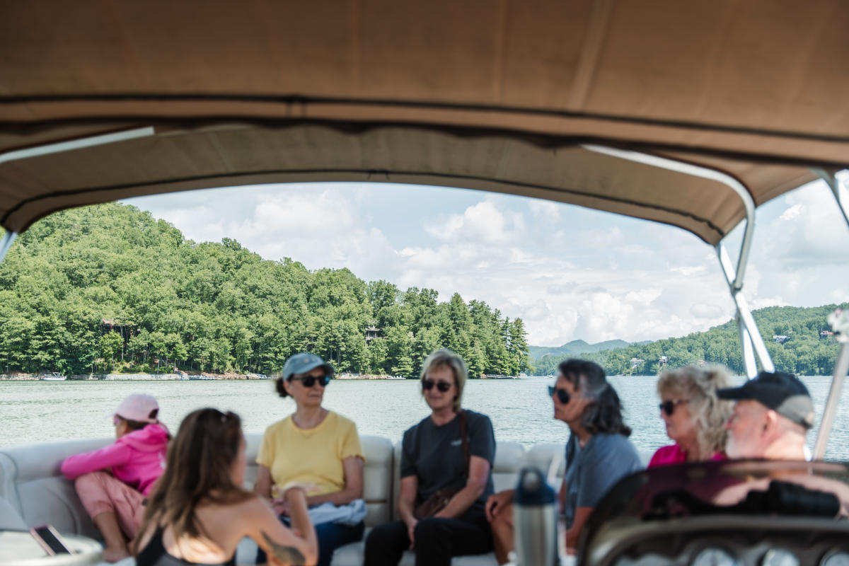 A group of people relaxing on a boat on a calm lake surrounded by lush green forested hills under a bright sky.