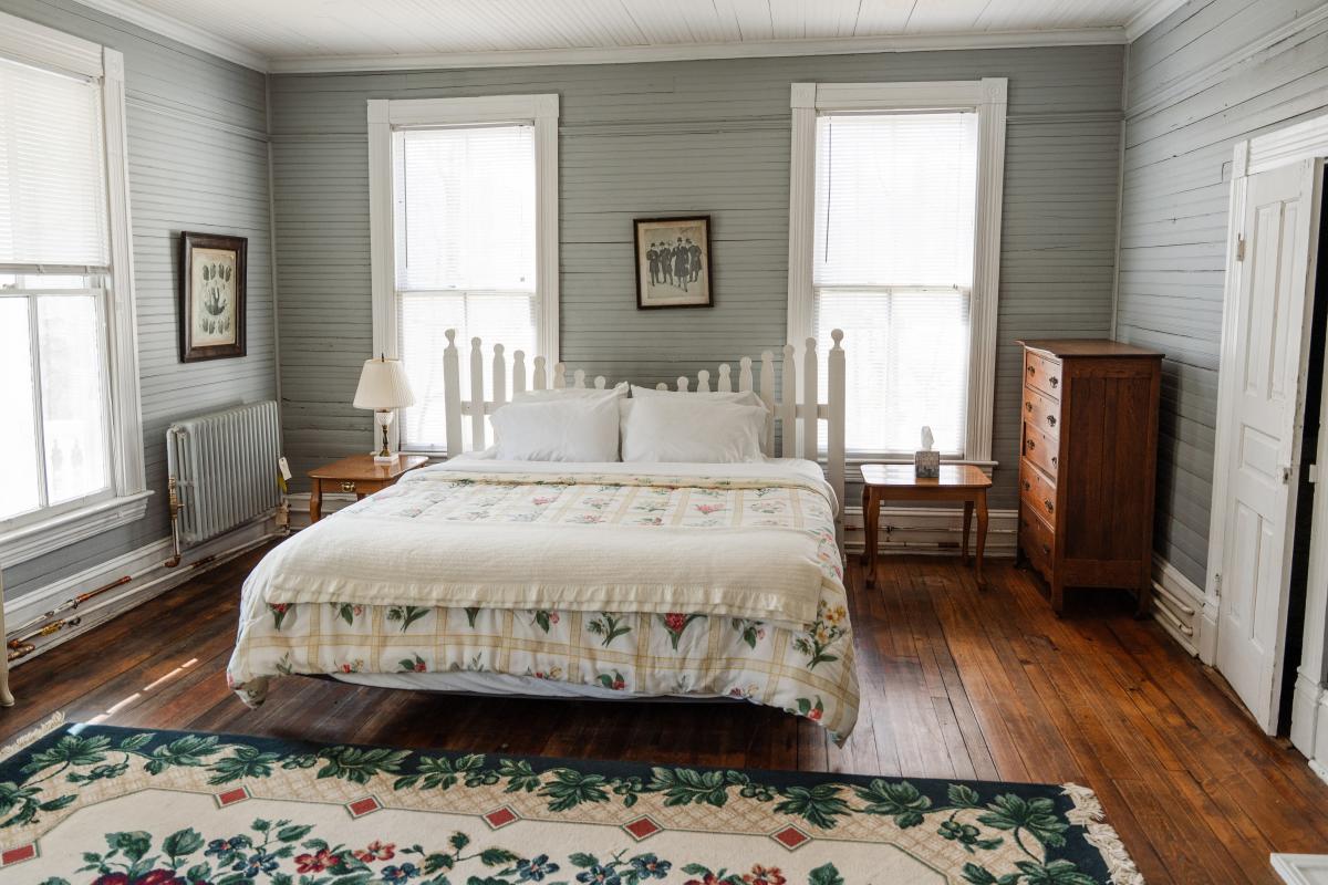 A wide shot of a rustic bedroom with gray-painted wood-paneled walls and dark hardwood floors. The room features a bed with a white picket-fence headboard and a floral quilt, a large decorative floral rug in the foreground, and a tall wooden dresser against the far wall.