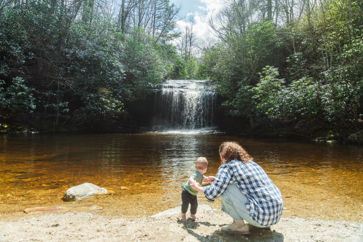 An adult with curly hair, wearing a blue and white plaid shirt, squats on a sandy shore holding the hands of a barefoot toddler in a green shirt. They are facing away from the camera, looking across a shallow, amber-colored pool. In the background, a wide, picturesque waterfall flows over a rock ledge, flanked by lush green trees and rhododendron bushes under a bright sky.