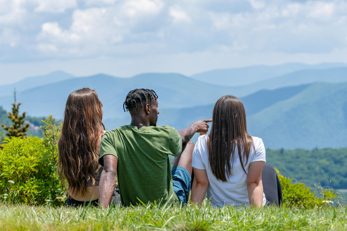 Three people sit together in lush green grass, viewed from behind, looking out over a vast mountain landscape. The person in the middle, wearing a green t-shirt, has their arm raised, pointing toward the distant, overlapping blue mountain ridges that stretch across the horizon under a bright, partly cloudy sky.