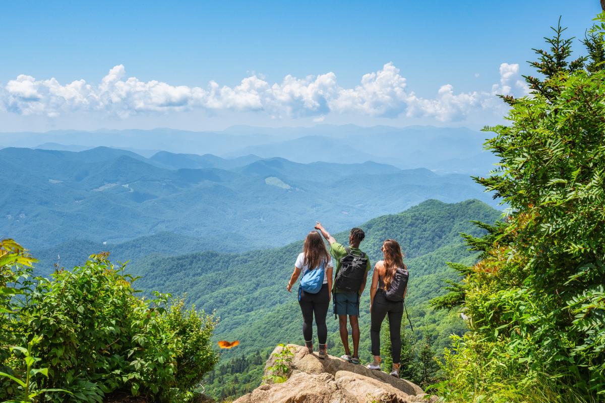 Three hikers with backpacks stand on a rocky ledge, looking out over a vast, hazy panorama of blue mountain ridges. One hiker points toward the horizon under a bright blue sky filled with fluffy white clouds.