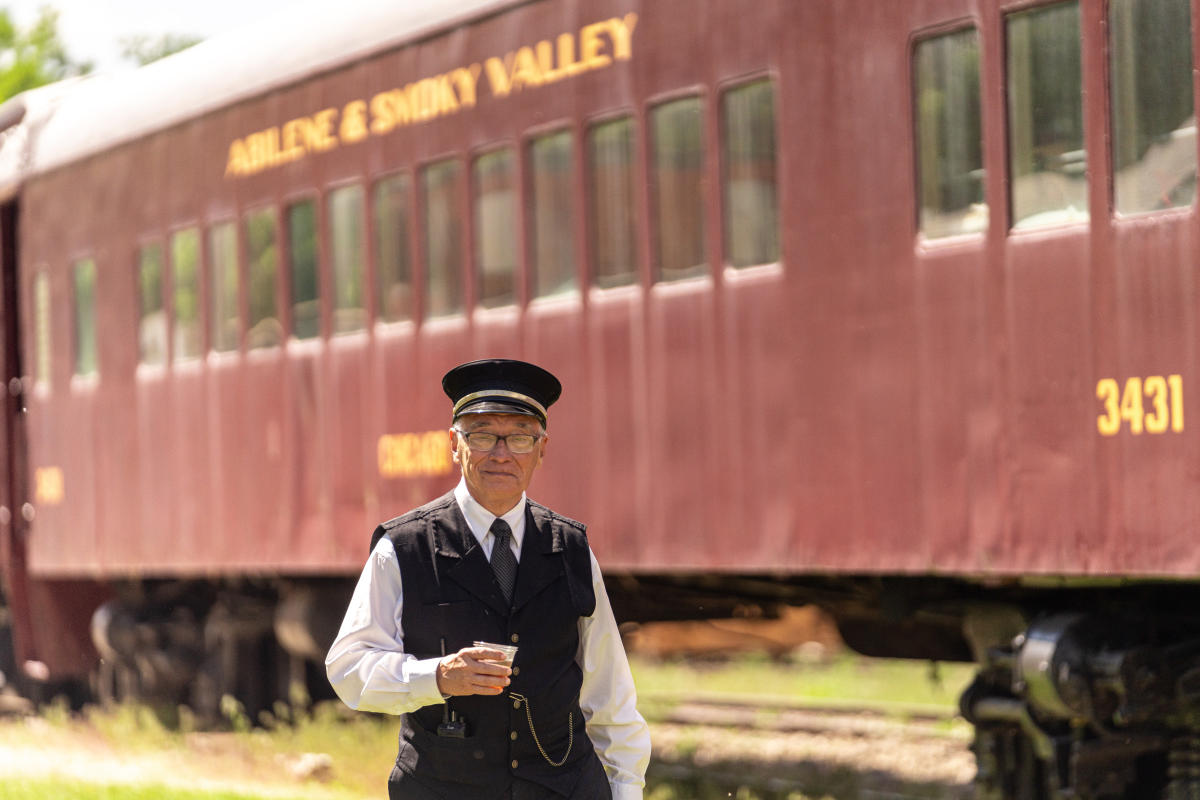 man, dressed as a historic train ticket taker, stands in front of a train holding a cup