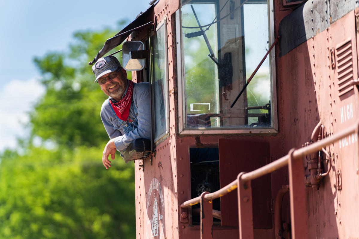a man in a train engineer uniform sticks his head out of a train window. He is smiling at the camera