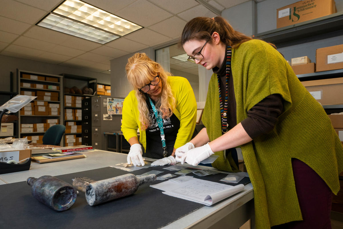 two women wearing white archival gloves look over historic artifacts on a table