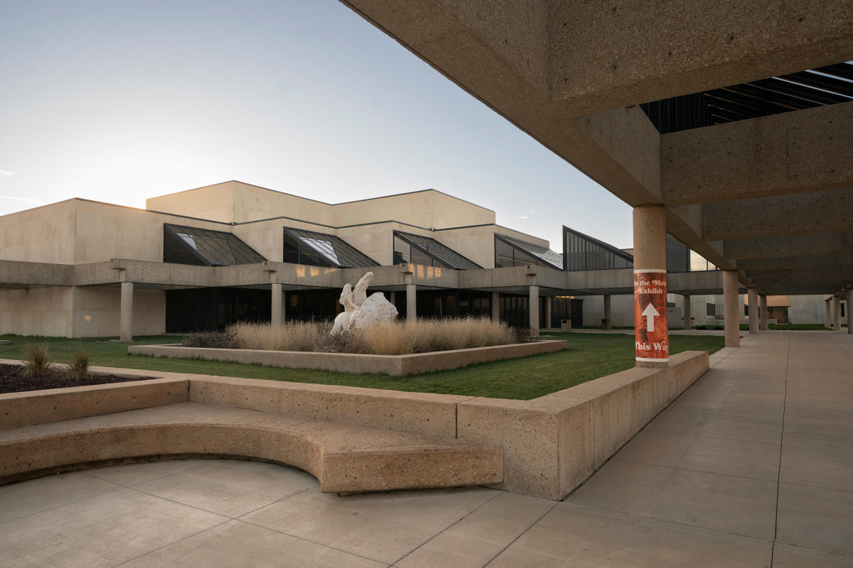 a geometric building with a side walk running along the building. A white statue of a Native person on a horses is surrounded by tall grass.