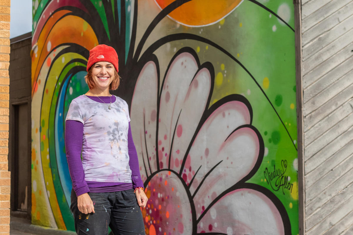 a woman wearing a red beanie and paint splattered clothes, stands in front of a street mural
