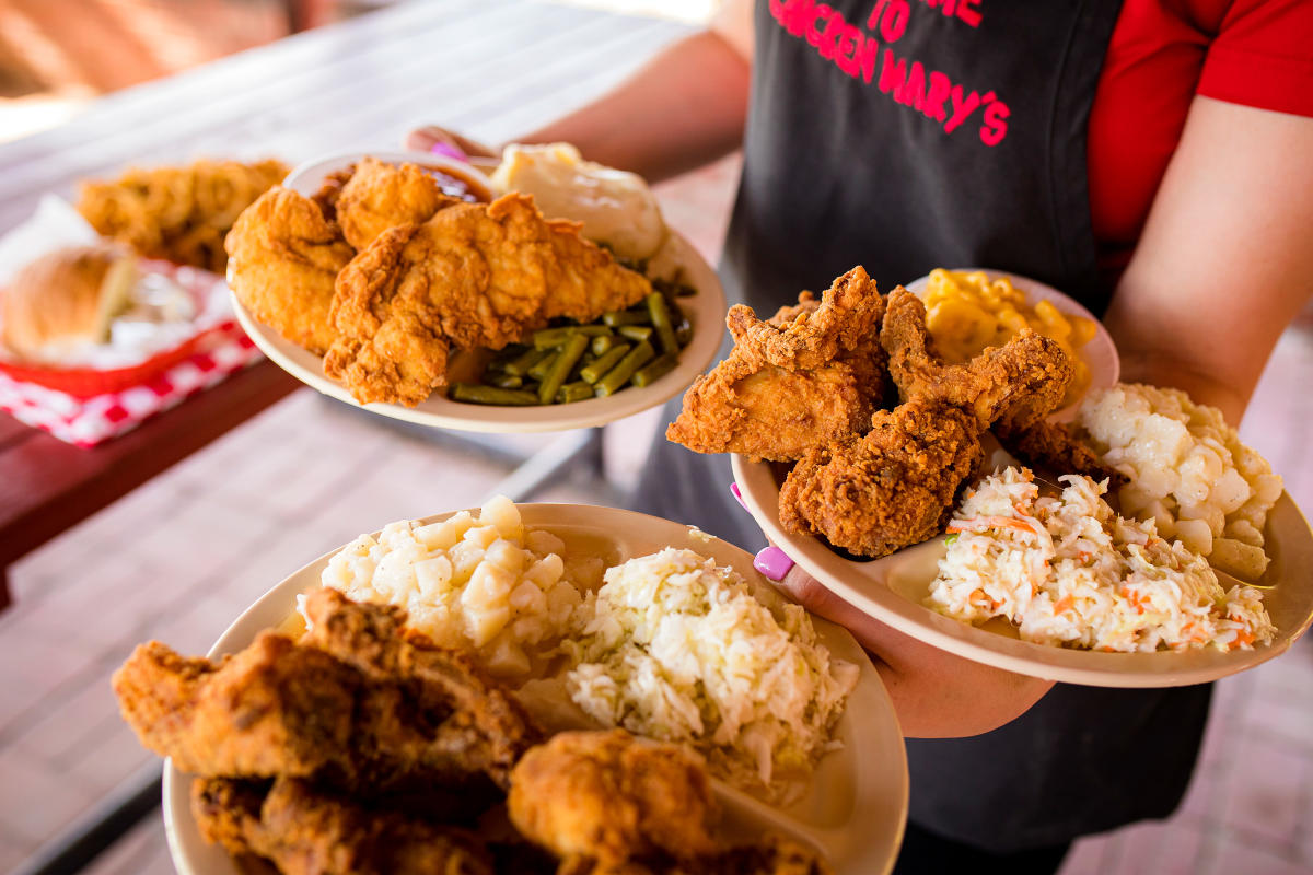 a waitstaff in a red t-shirt and black apron, is carrying three plates of fried chicken and sides.