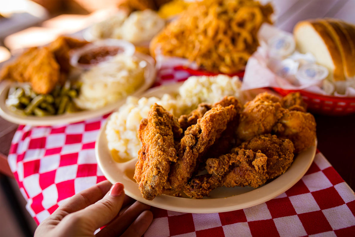 A woman's hand grabs a plate of fried chicken and sides off a table with a red and white checkered table cloth. In the background, there are additional plates of fried chicken.