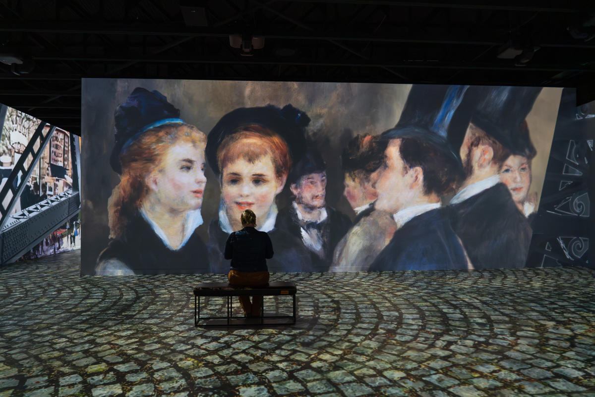 a woman sits on a bench looking a digital painting projected on to a wall